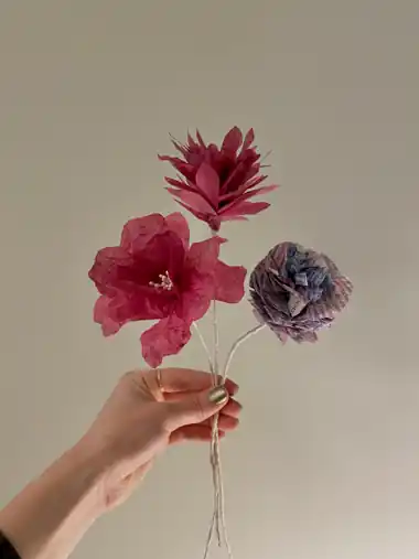 Drei Krepppapierblumen in Rot, Bordeaux und Violett mit Schriftmuster, von einer Hand gehalten