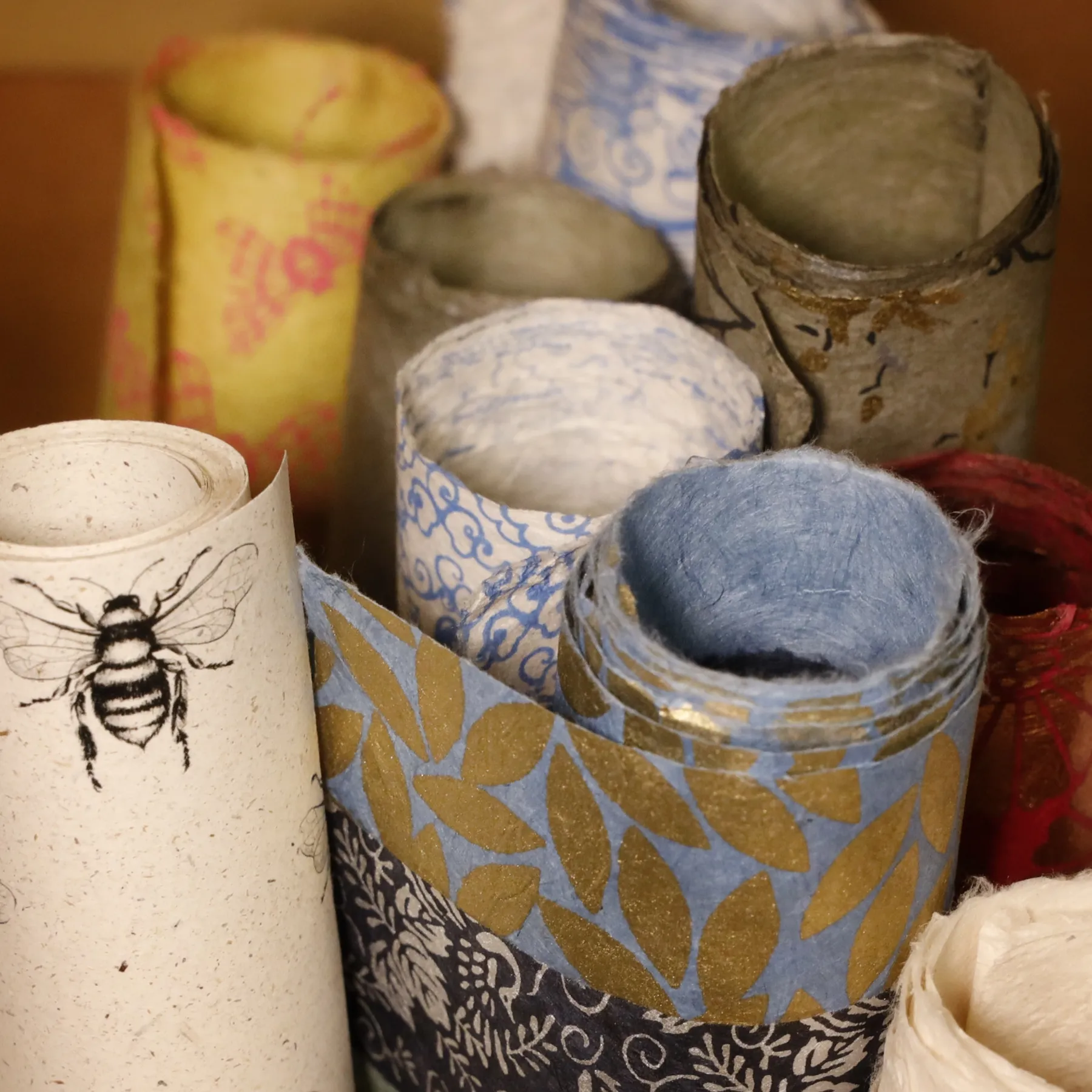 Aufgerollte Geschenk- und Musterpapiere in Blau, Gold und Weiß, darunter ein Papier mit Bienenmotiv