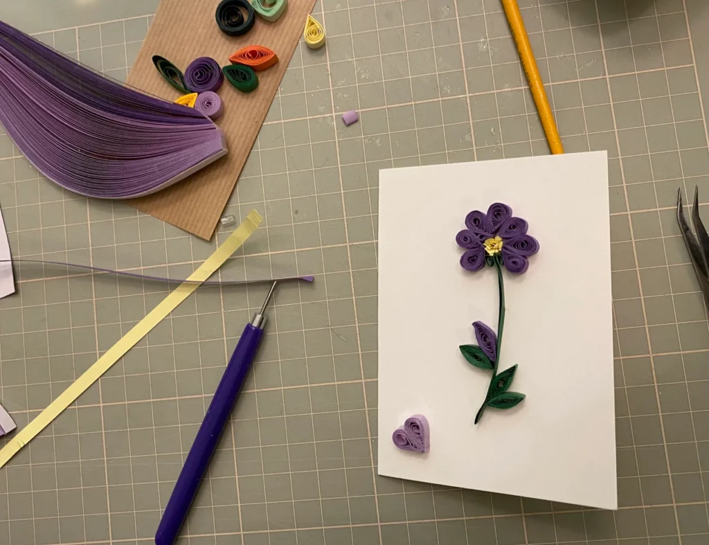 Quilling-Karte mit violetter Blume aus gerollten Papierstreifen auf einer Schneidematte