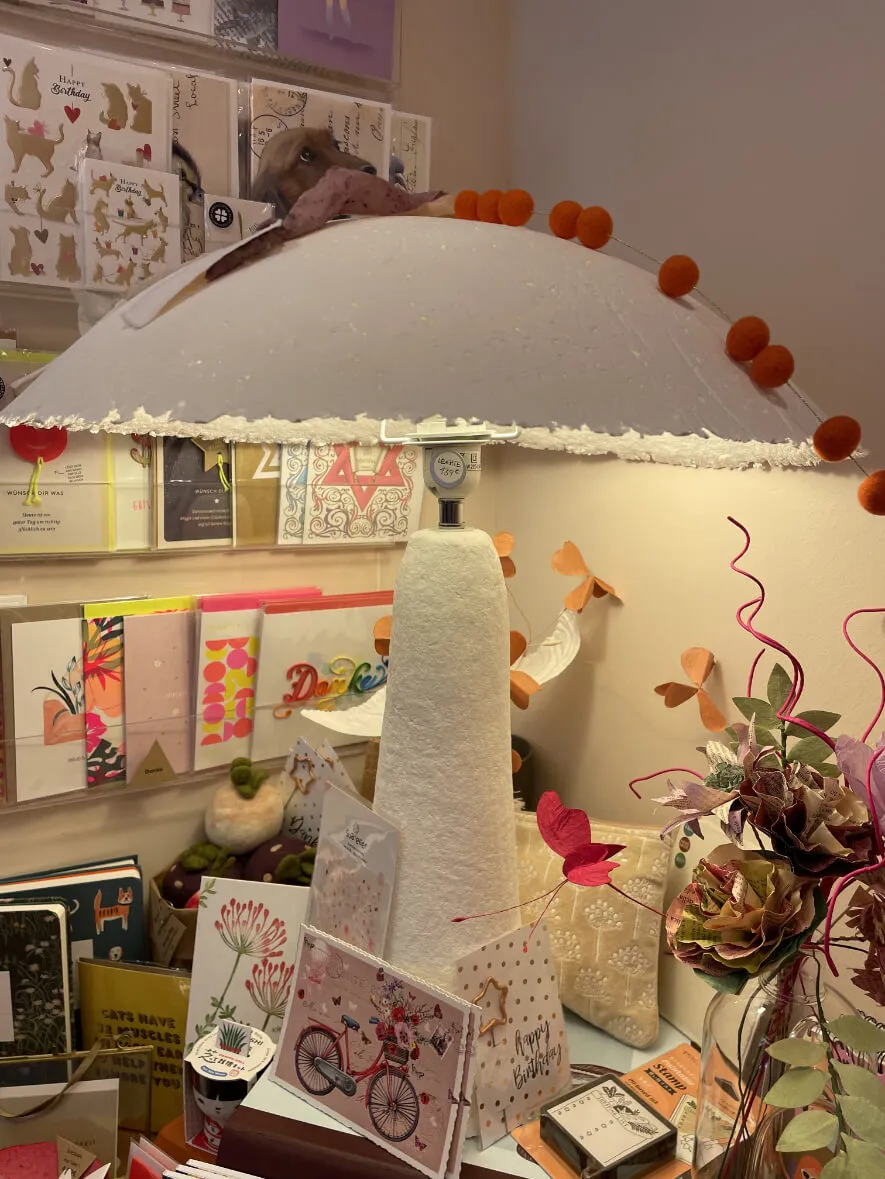 Pilzförmiger Papierschirm mit orangefarbenen Pompons vor einem Regal mit Karten