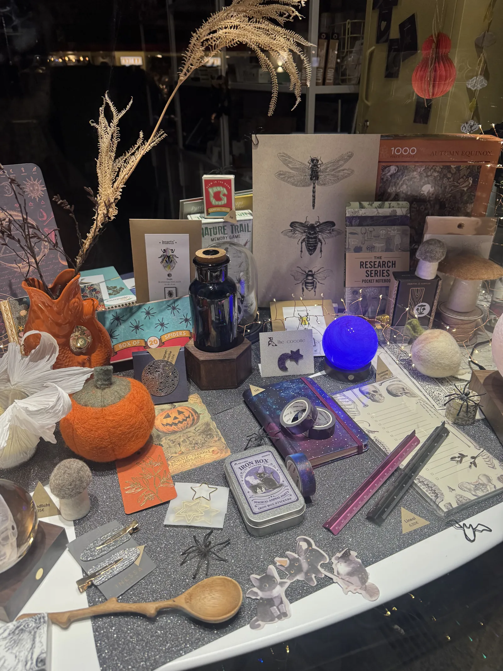Halloween-Schaufenster von L' Atelier Papier: Filzkürbis, Libellen-Poster, blaue Leuchtkugel, Notizbücher und Papeterie auf glitzerndem Untergrund