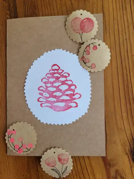 Kraftpapier-Weihnachtskarte mit rosa gestempeltem Tannenzapfen und gestempelten runden Etiketten auf Holztisch